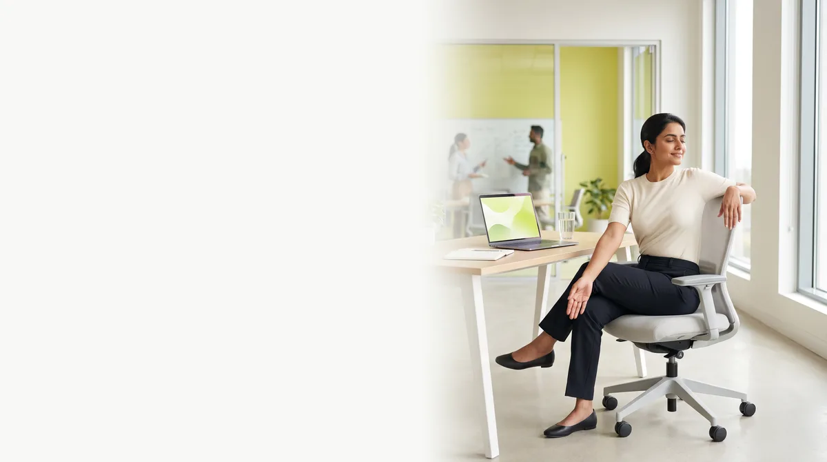 An Indian professional doing a seated yoga twist on her office chair at her desk, a learning course open on her laptop, colleagues at a whiteboard behind her